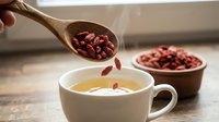 Hand pouring red adzuki beans from wooden spoon into herbal tea cup on rustic kitchen counter. Healthy organic legumes preparation for cooking.