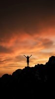 Silhouette of person with raised arms celebrating on rocky mountain peak against dramatic orange sunset sky with clouds for success and achievement concepts.