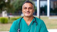 Middle-aged Hispanic male doctor in green scrubs with stethoscope smiling confidently outside modern medical facility for healthcare marketing.