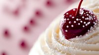 Fresh cherry with silver sugar pearls on top of creamy white frosting swirl, macro food photography with soft pink bokeh background creates elegant dessert scene.