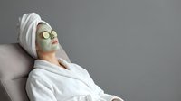 Young woman relaxing in white bathrobe with cucumber eye mask and towel on head during spa treatment for skincare and wellness routine.