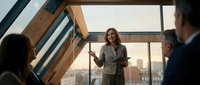Professional businesswoman presenting to colleagues in modern office with wooden beams and city skyline view during corporate meeting or training session.