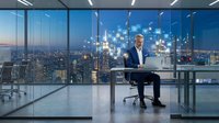Caucasian middle-aged businessman in blue suit working on laptop in modern office with city skyline and glowing digital icons at night.