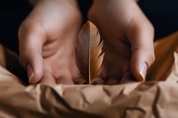 Female hands gently holding brown feather over crumpled kraft paper background in warm tones, creating minimalist composition with soft natural lighting and shallow depth of field.