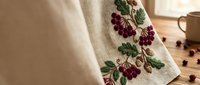 Linen kitchen towel with embroidered grape vine pattern on wooden table with coffee beans and mug creating cozy rustic home decor atmosphere.