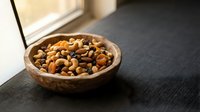 Mixed nuts and dried fruits in wooden bowl on dark surface near window with natural lighting for healthy snacking and nutrition concepts.