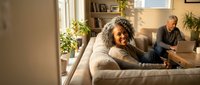 Smiling African American senior woman relaxing on couch at home with husband reading in background. Comfortable retirement lifestyle indoors.