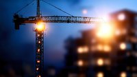 Construction crane silhouette against dramatic evening sky with blurred residential building and glowing lights creating atmospheric urban development scene.