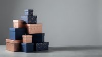Stack of decorative gift boxes in copper, navy blue and patterned designs arranged in pyramid formation against neutral background for packaging concepts.