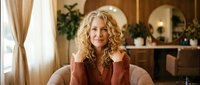 Professional blonde woman with curly hair sitting confidently in modern office interior, wearing brown blazer for business lifestyle content.