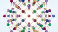 Young woman with long brown hair and fair complexion, wearing casual clothing, smiling while holding a colorful molecular structure model with interconnected spheres and rods in purple, blue, green, and gold tones against white background.