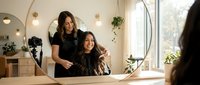 Young female hairstylist styling brunette woman's hair in modern salon with natural lighting and plants creating warm professional atmosphere.