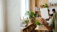 Person with medium skin tone holding blank white card in modern home office with wooden shelves, potted plants, and natural window light, suitable for business messaging and lifestyle marketing.