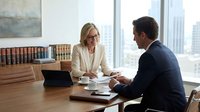 Woman in business casual attire and glasses discusses documents with man in dark suit at wooden desk in modern office with city skyline view.