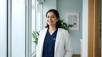Young South Asian female healthcare professional wearing white lab coat and navy scrubs, standing in modern medical office with natural light from windows and green plants, suitable for healthcare, medical practice, hospital, and wellness industry marketing materials.
