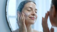 Young woman with South Asian ethnicity applying moisturizer or skincare product to her face while looking in mirror. Ideal for beauty, cosmetics, skincare routines, wellness and personal care advertising campaigns.