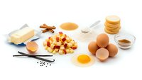 Fresh baking ingredients arranged on white background including eggs, butter, flour, vanilla, cinnamon and diced apples for homemade dessert preparation.