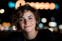 Young woman with short wavy hair smiling at camera against blurred city lights bokeh background at night. Urban lifestyle portrait for social media content.