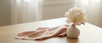 Cream peony flower in white ceramic vase on wooden table with blush pink fabric, soft natural light, minimalist home decor styling.