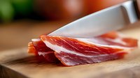 Thinly sliced prosciutto ham on wooden cutting board with chef knife, macro shot showing delicate meat texture and marbling. Fresh gourmet Italian cured meat.