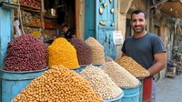 Middle Eastern bearded merchant smiling at traditional spice market stall with colorful heaps of nuts, legumes and dried goods displayed in large bins.