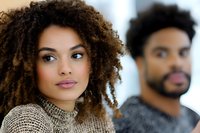 Young African American woman with natural curly hair and confident expression wearing beige turtleneck sweater, with male figure blurred in background. Professional portrait.