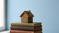 Small wooden house model sitting on stack of vintage books near window symbolizing home education and knowledge foundation for real estate investment.
