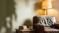 Cozy living room corner with brown leather armchair, soft throw blanket, wooden side table with books and coffee mug, warm table lamp creating inviting atmosphere.