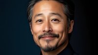 Middle-aged Asian man with gray-streaked hair and facial hair smiling warmly against dark background, professional headshot style portrait showing confidence and approachability.