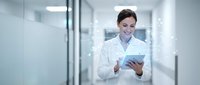 Young female doctor in white coat using digital tablet while walking through modern hospital corridor with bright lighting and glass walls.