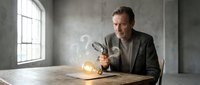Middle-aged Caucasian businessman in gray suit examining glowing light bulb with magnifying glass at wooden desk in modern office interior with concrete walls.