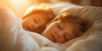 Two young Caucasian children sleeping peacefully in bed with warm golden sunlight streaming through window, creating serene morning atmosphere.
