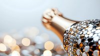 Luxurious champagne bottle decorated with silver sequins and sparkles against blurred bokeh lights background creating festive celebration atmosphere.