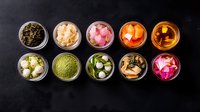 Colorful assortment of Asian pickled vegetables and condiments in glass jars viewed from above on dark background, including ginger, radish and seaweed.