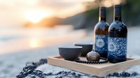 Wine bottles with blue floral patterns and ceramic bowl on wooden tray at sunset beach setting, creating romantic coastal atmosphere with soft golden light.