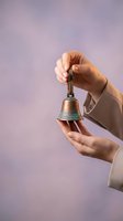 Caucasian woman hands holding antique bronze bell with patina against soft purple background, symbolizing announcement or celebration.