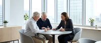 Three professionals in business attire discuss documents at a modern office table with city skyline views through floor-to-ceiling windows, suitable for corporate, teamwork, and business meeting concepts.