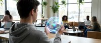 Young man examining holographic globe projection in modern coworking space with diverse professionals working at desks in bright office environment.