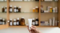 Mature woman's hand holding blank white cosmetic tube against shelves of skincare products and bottles in modern wellness store or spa setting.