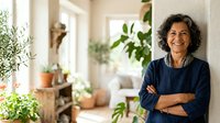 Mature woman with curly dark hair and medium skin tone, wearing a dark navy button-up shirt, standing with arms crossed in a bright, plant-filled home interior. Potted plants and natural light create a welcoming, botanical atmosphere for lifestyle and wellness content.