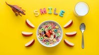 Colorful cereal bowl with fruit loops, strawberries, apple slices, milk, and SMILE letters on bright yellow background, fun kids breakfast concept.