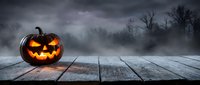 Glowing jack-o-lantern pumpkin with carved face sits on wooden deck against misty forest backdrop. Halloween decoration for seasonal marketing campaigns.