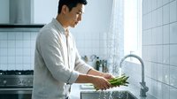 Asian man washing fresh green asparagus vegetables under running water in modern kitchen sink for healthy cooking preparation at home.