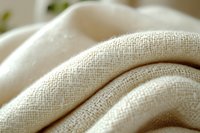 Soft folds of natural linen fabric with visible textile texture and weave pattern in warm beige tones, macro photography showing detailed cloth structure.