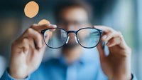 Person holding up modern eyeglasses with black frames against blurred background, demonstrating proper lens cleaning technique and eyewear maintenance.
