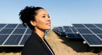 Woman with dark curly hair, South Asian ethnicity, wearing black blazer, smiling while standing in front of solar panels on sunny day. Renewable energy professional.