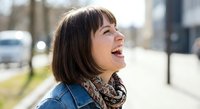 Happy young Caucasian woman with brown bob haircut laughing outdoors in denim jacket and patterned scarf on sunny day in urban setting.