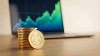 Stacked gold coins with cryptocurrency symbol leaning against pile on wooden desk, laptop displaying rising green financial chart in background for investment and trading concepts.
