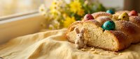 Freshly baked Easter bread with colorful eggs and a small bunny figurine on a yellow linen cloth in warm sunlight.