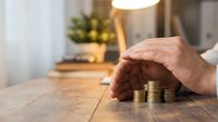 Protective hands covering stack of golden coins on wooden table in cozy home interior representing financial security and savings protection concept.